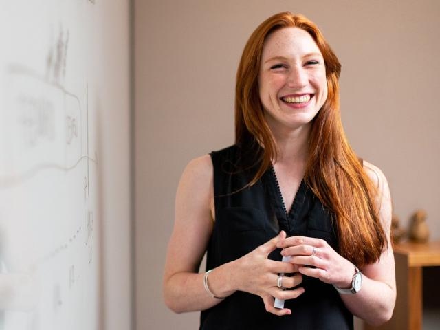 Smiling woman with long hair wearing a black dress and standing in front of a whiteboard